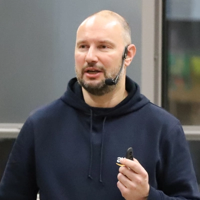 Alexander Günsche speaking with a headset microphone, wearing a dark blue hoodie and holding a presentation remote.